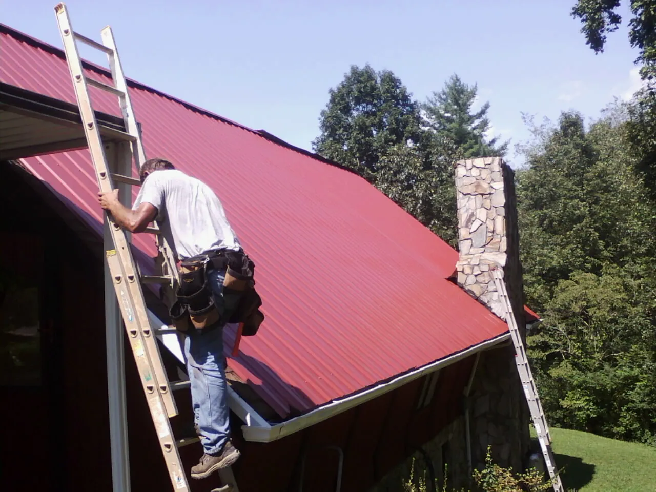 Roofer on a ladder beside a red metal roof and stone chimney — Soffit & Fascia Repair by Roofing Brothers in Menomonie